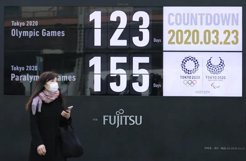 3月23日，一名行人戴着口罩在日本东京新桥地区经过显示东京奥运会和残奥会倒计时的大屏幕。图片来源：新华社